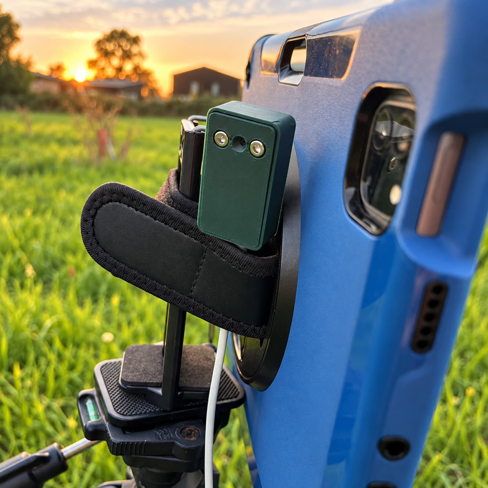 The dark green bat dongle mounted beside a compact field recorder on a tripod, in a grassy field at sunset.