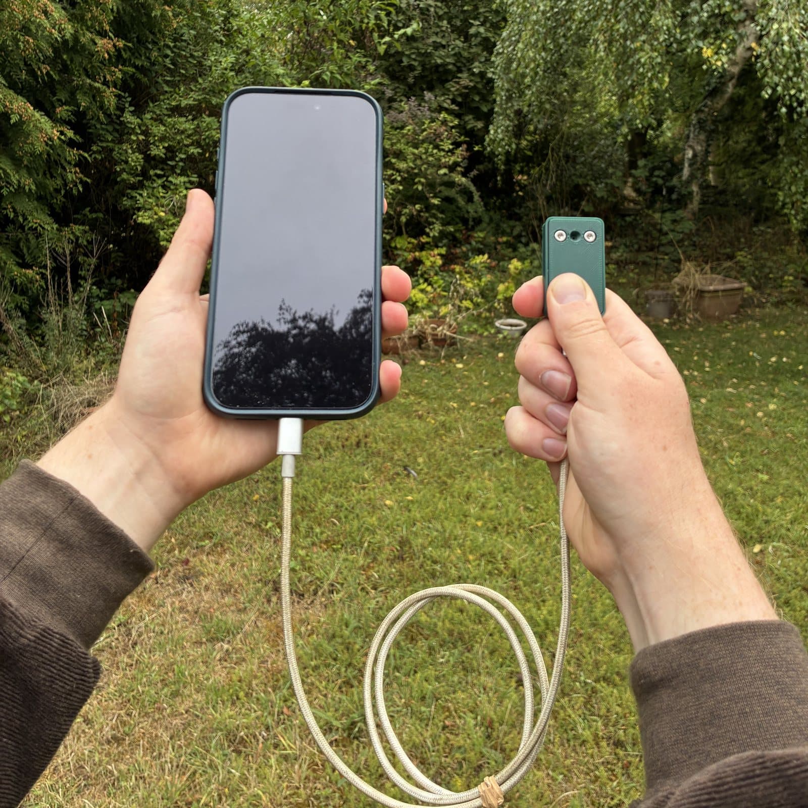 Two hands outdoors in a garden, one holding a phone and the other holding the bat dongle up, connected by a looped USB-C cable.