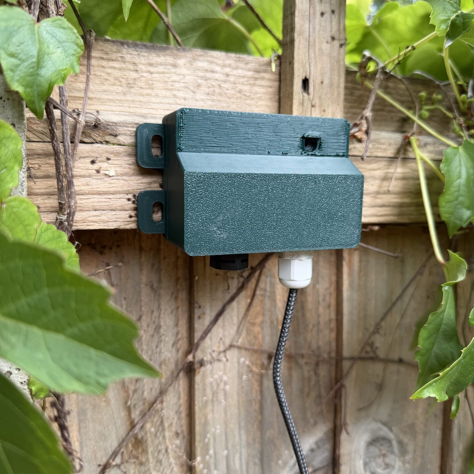 The alauda listening post mounted on a garden fence rail, seen from three-quarters, with a cable running from the gland on its underside.